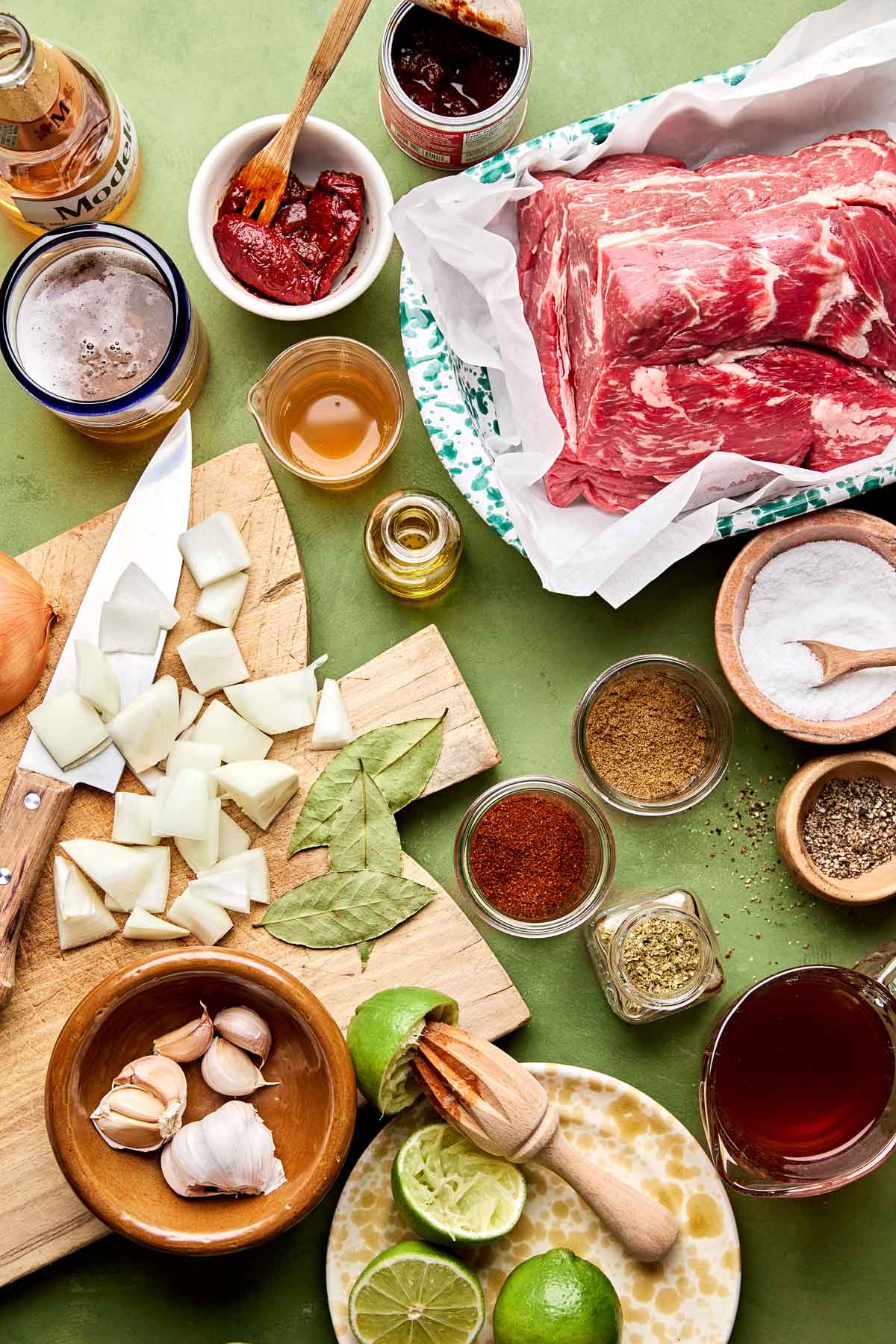 An overhead shot of ingredients displayed on a wooden cutting board and in various dishes on a green surface: beef chuck roast, salt and pepper, onions, bay leaves, garlic, limes, chipotle peppers, beer, apple cider vinegar, chili powder, cumin, oregano, beef stock, and olive oil.