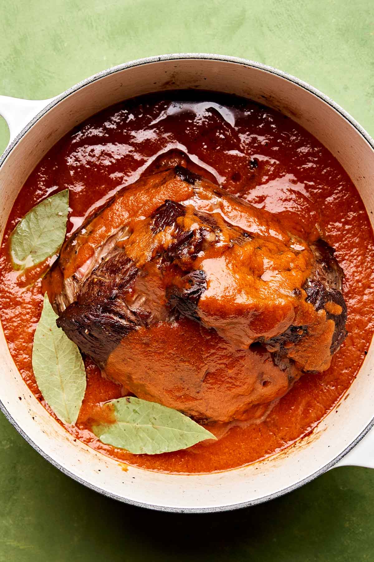 An overhead shot of chuck roast in braising liquid with three bay leaves set on top in a large white Dutch oven atop a green surface.