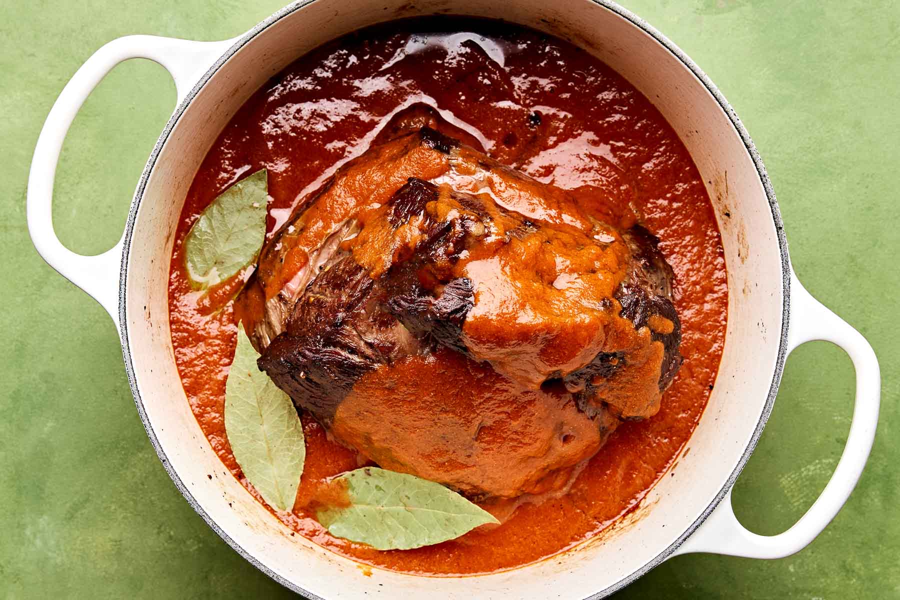An overhead shot of chuck roast in braising liquid with three bay leaves set on top in a large white Dutch oven atop a green surface.