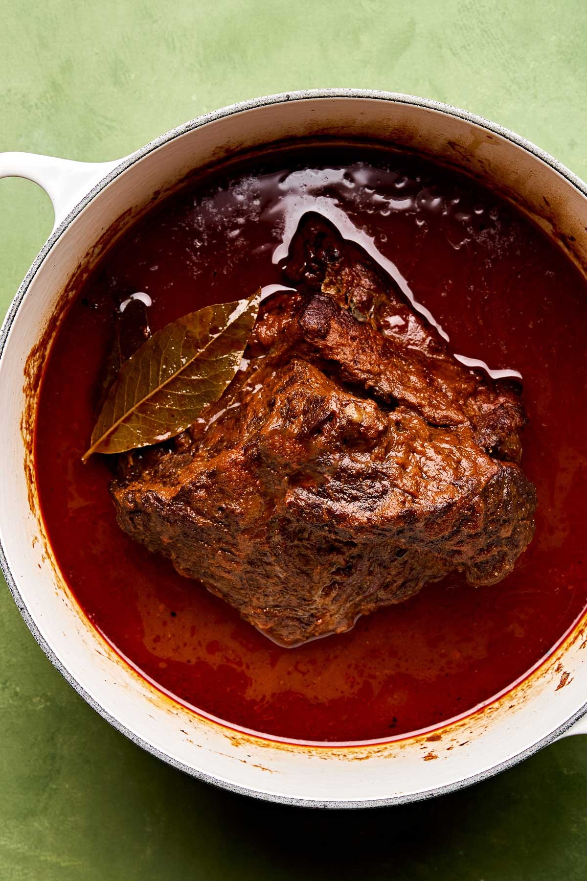 An overhead shot of braised beef chuck roast in its braising liquid in a large white Dutch oven atop a green surface.