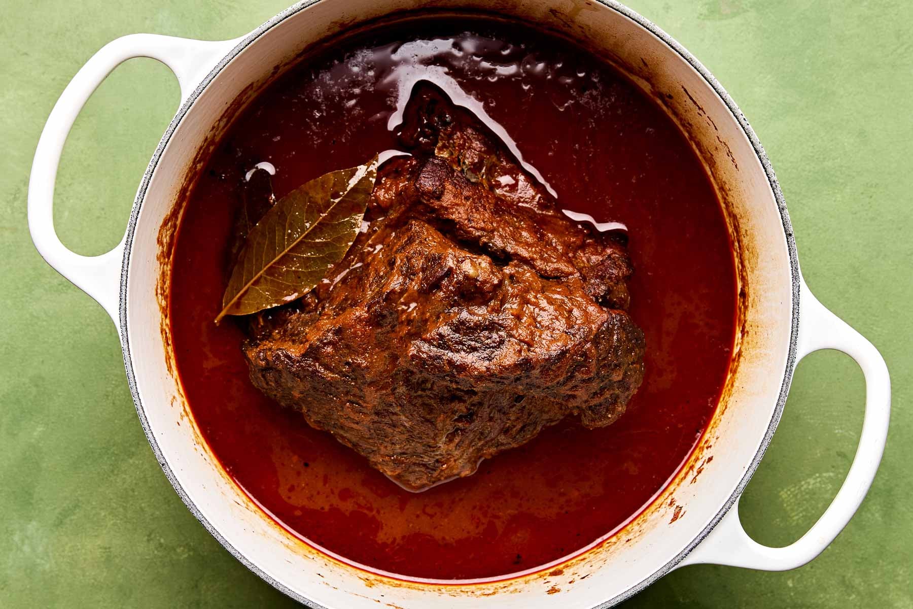 An overhead shot of braised beef chuck roast in its braising liquid in a large white Dutch oven atop a green surface.