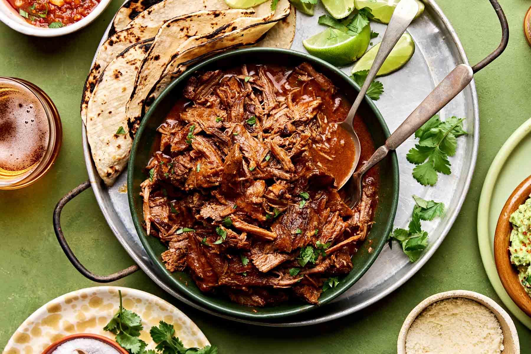 An overhead shot of braised beef chuck roast taco meat in a large green bowl alongside charred corn tortillas and lime wedges on a round tin tray. The tray sits on a green surface surrounded by dishes of cotija cheese, salsa, beer, salt, cilantro and guacamole.