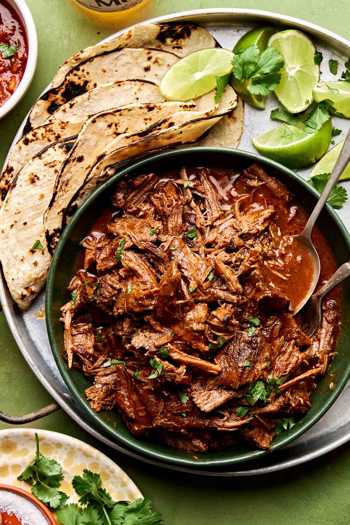 An overhead shot of braised beef chuck roast taco meat in a large green bowl alongside charred corn tortillas and lime wedges on a round tin tray. The tray sits on a green surface surrounded by dishes of cotija cheese, salsa, beer, salt, cilantro and guacamole.