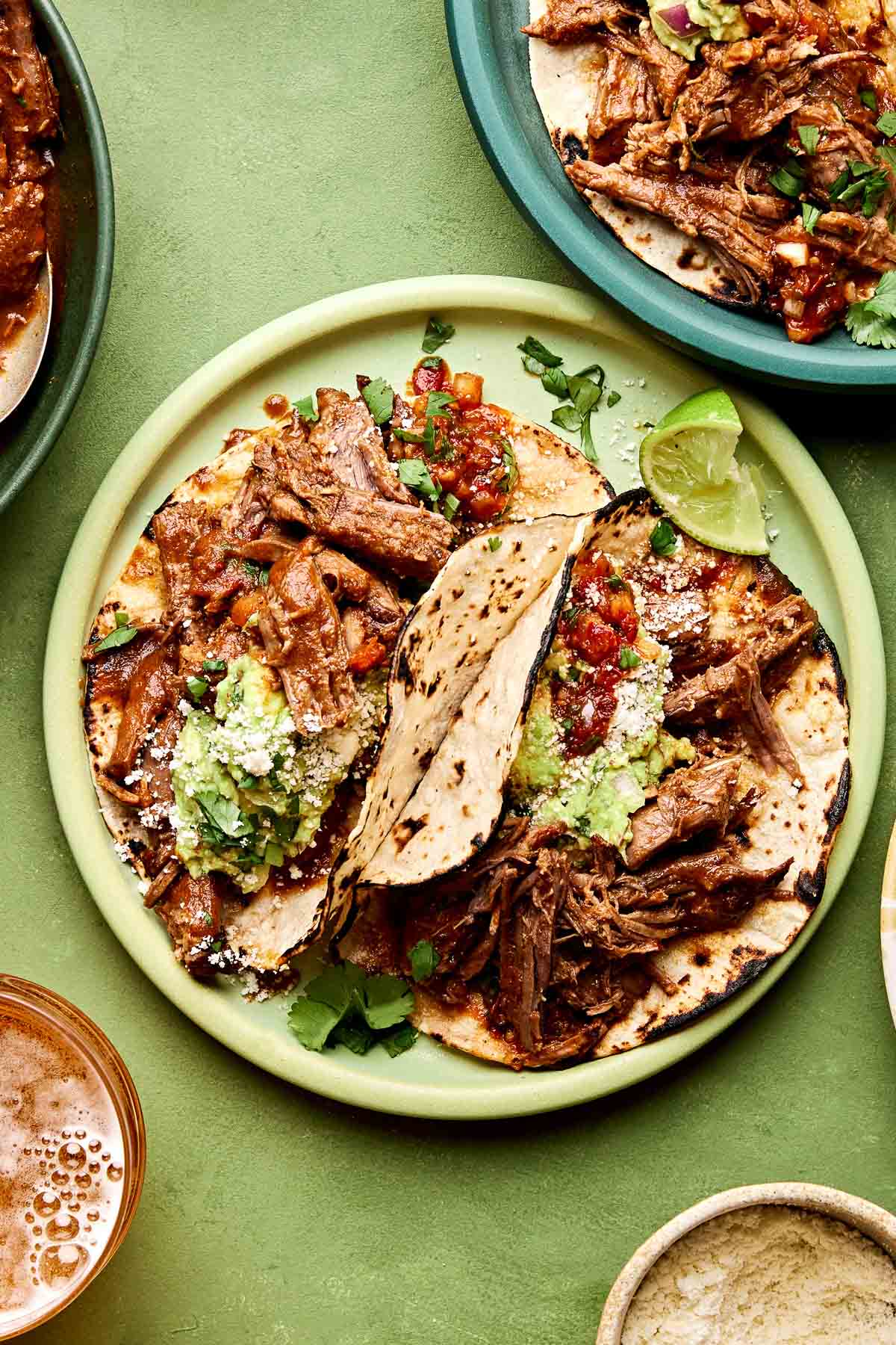An overhead shot of two beef chuck roast tacos topped with salsa, cotija cheese and guacamole on charred corn tortillas on a lime green plate. The plate sits on a green surface alongside another dish of tacos, a cup of beer and a dish of cheese.
