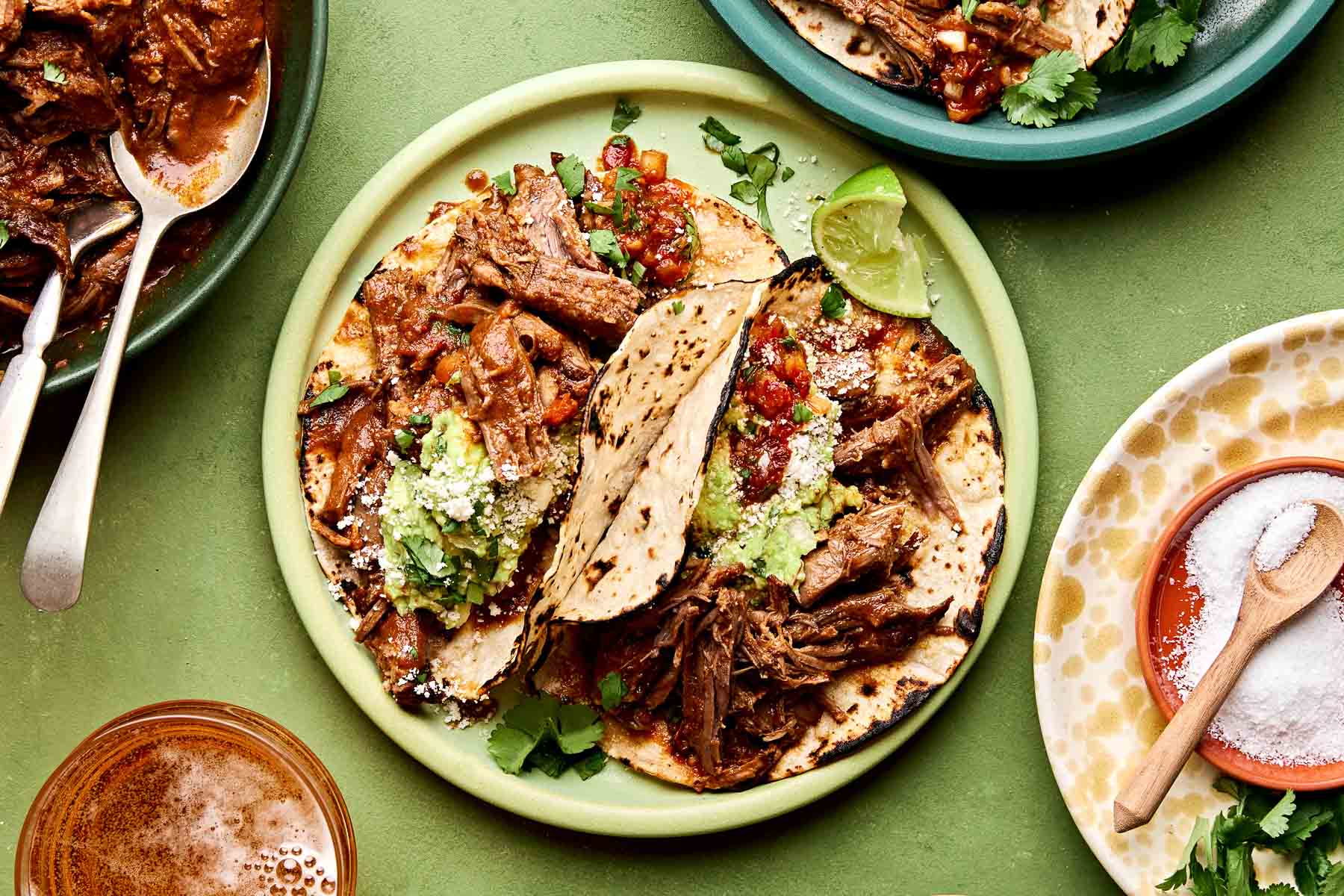 An overhead shot of two beef chuck roast tacos topped with salsa, cotija cheese and guacamole on charred corn tortillas on a lime green plate. The plate sits on a green surface alongside another dish of tacos, a cup of beer and a dish of cilantro and salt.