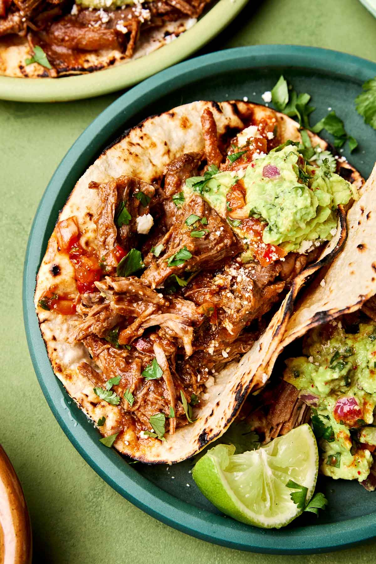 A close-up angled shot of two shredded beef chuck roast tacos on a teal plate atop a green surface.