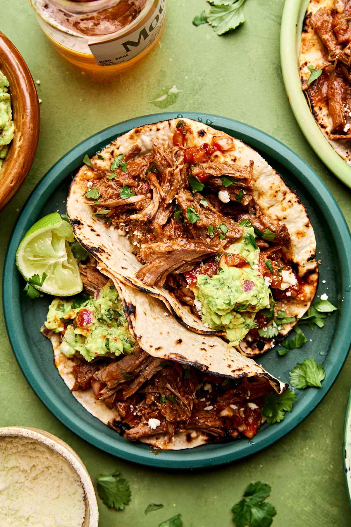 An overhead shot of two beef chuck roast tacos topped with salsa, cotija cheese and guacamole on charred corn tortillas on a teal plate. The plate sits on a green surface alongside another dish of tacos, a bottle of beer and a dish of cheese.