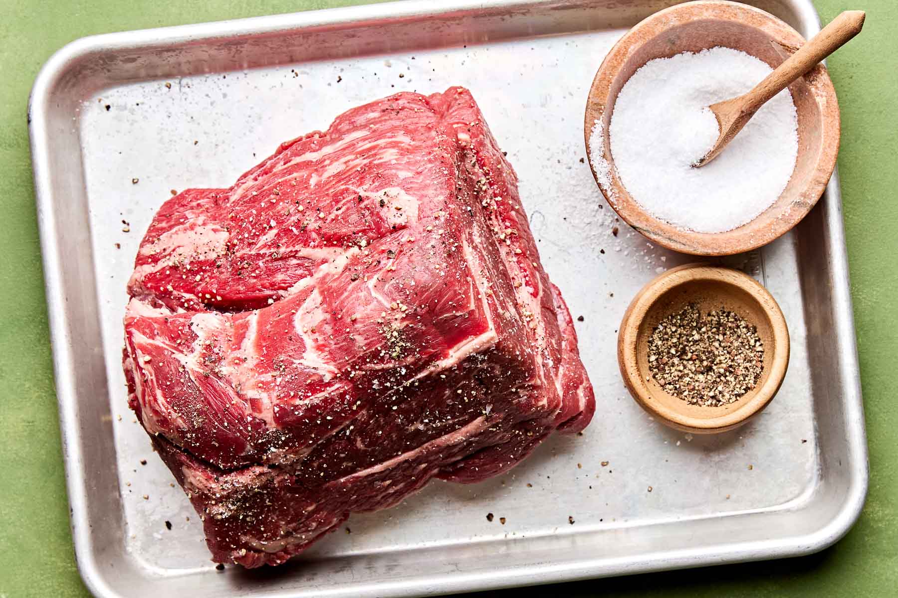 An overhead shot of chuck roast seasoned with salt and pepper on a small sheet pan atop a green surface. Small dishes of salt and pepper sit alongside the roast.