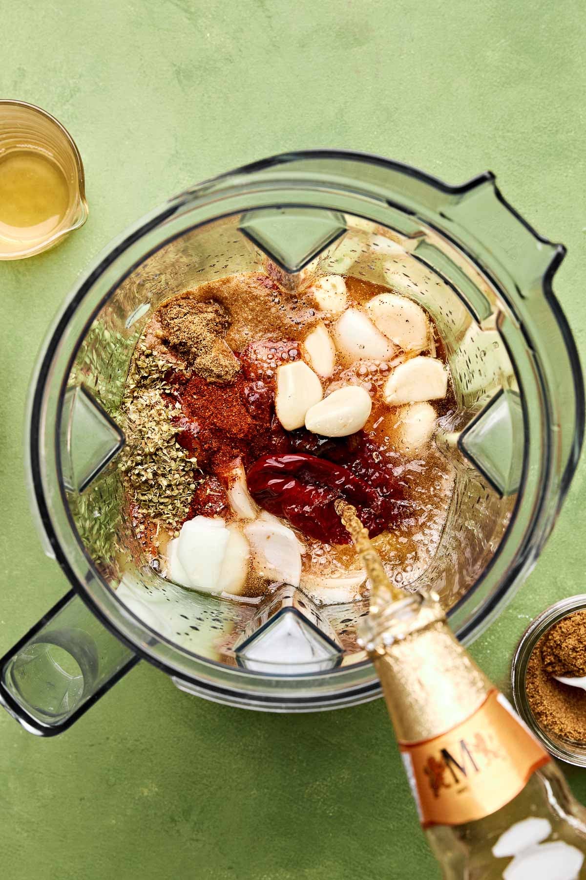 An overhead shot of beer being poured over spices, garlic, onions, and chipotle peppers in a blender sitting atop a green surface.