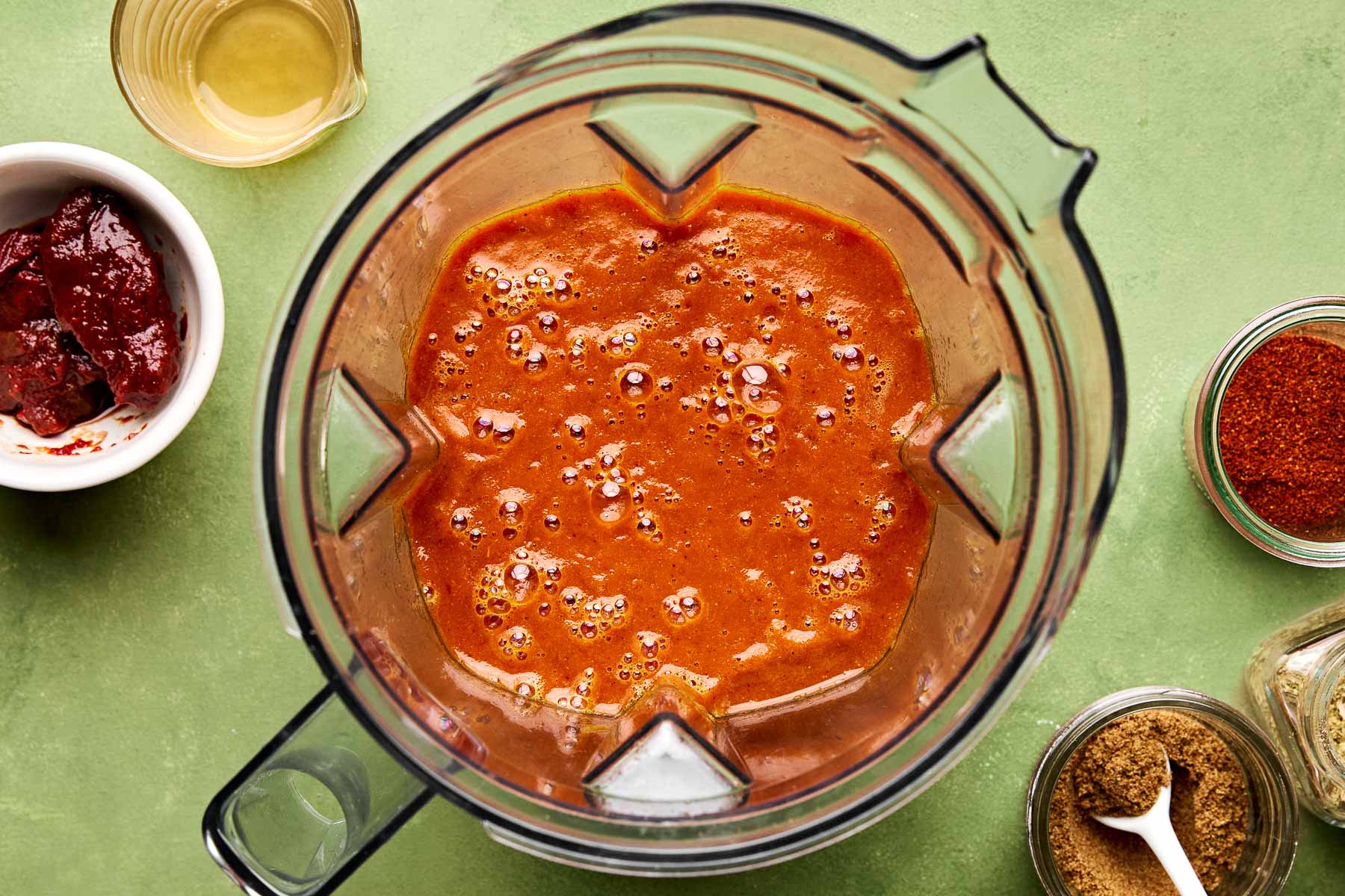 An overhead shot of blended braising liquid in a blender pitcher atop a green surface. The blender is surrounded by dishes of chipotle peppers, olive oil, and spices.