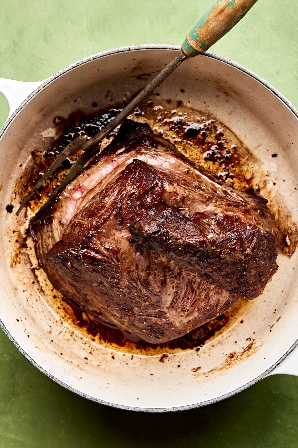 An overhead shot of browned beef chuck in a large Dutch oven atop a green surface.