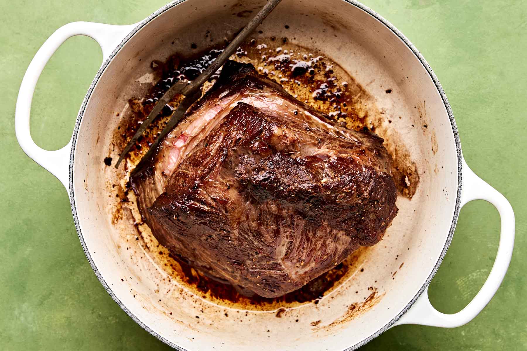 An overhead shot of browned beef chuck in a large Dutch oven atop a green surface.