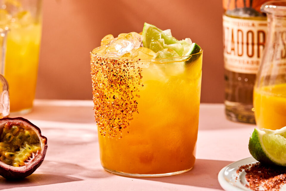 A close-up side shot of a lilikoi margarita in a tajin-rimmed glass garnished with a lime wedge. The glass sits on a light pink surface with a pitcher of margaritas and a bottle of tequila in the background. A dish of tajin and a lime wedge, a small pitcher of passion fruit puree, and half a passion fruit sit alongside the margarita.