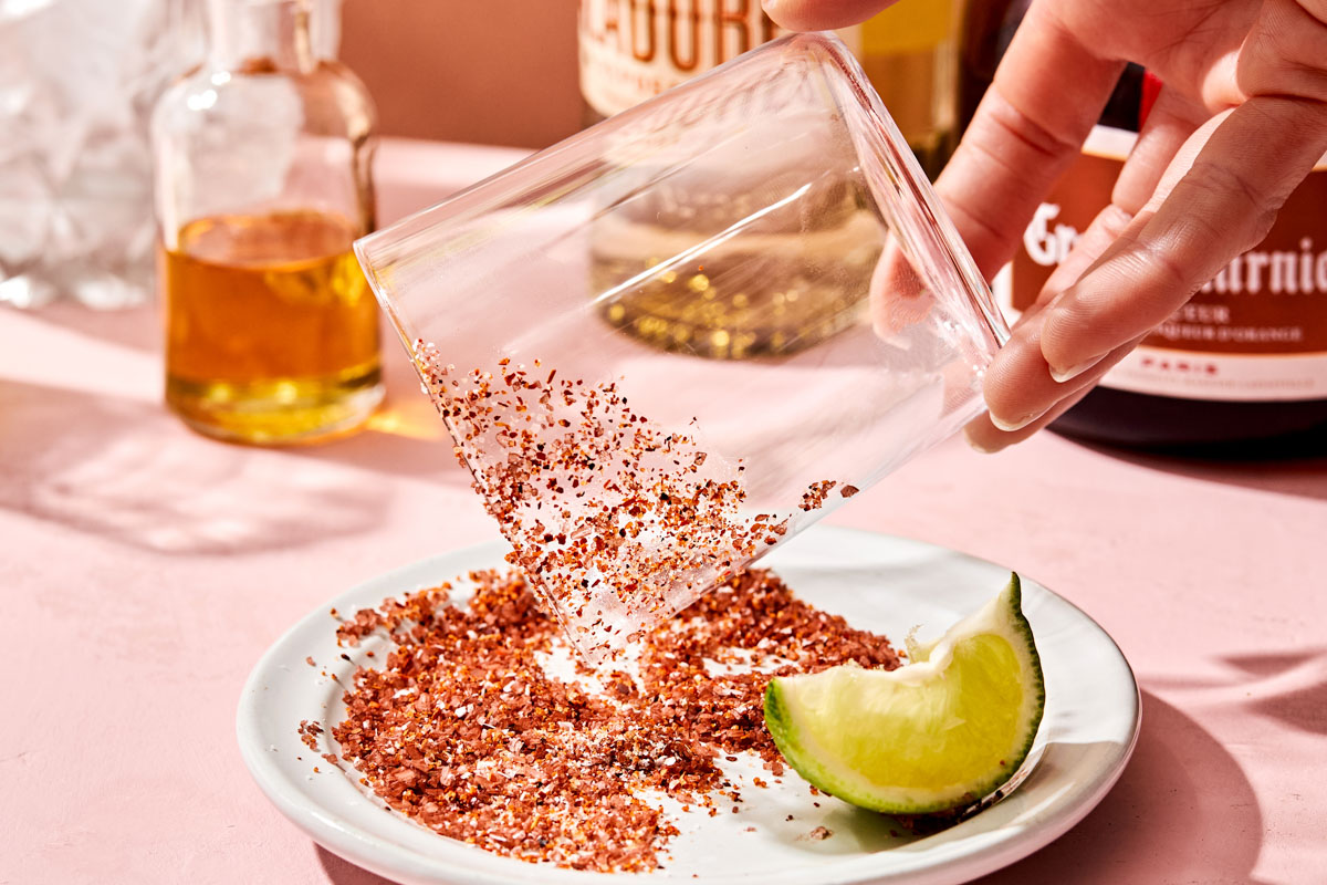 A side shot of a woman's hand rimming a glass with tajin seasoning. A white dish of tajin sits on a pink surface, and bottles of agave, tequila and Grand Marnier sit in the background.