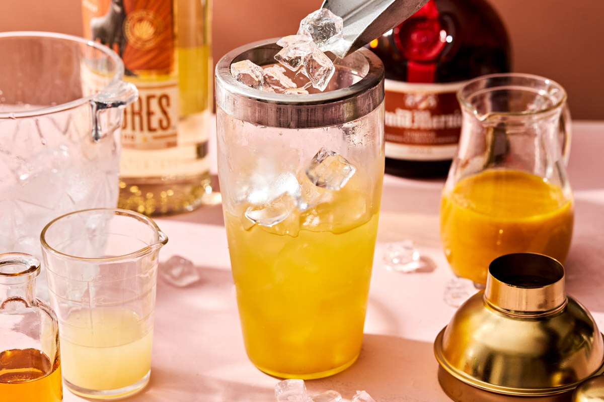 A side shot of a scoop dropping ice into a glass cocktail shaker with a silver rim. The shaker sits on a pink surface, with glasses of passion fruit puree and lime juice alongside it, and bottles of tequila and Grand Marnier in the background.