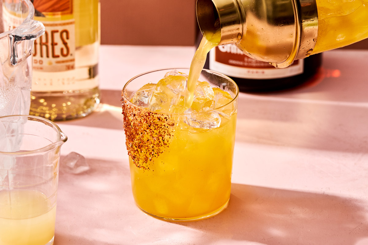 An angled side shot of a lilikoi margarita being poured from a cocktail shaker into a glass rimmed with tajin. The glass sits on a pink surface with bottles of tequila and Grand Marnier in the background.
