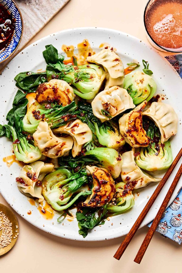 An overhead shot of cooked dumplings and bok choy drizzled with sauce on a white plate on a beige surface. A pair of chopsticks sits alongside the plate, along with dishes of sesame seeds and dumpling sauce.