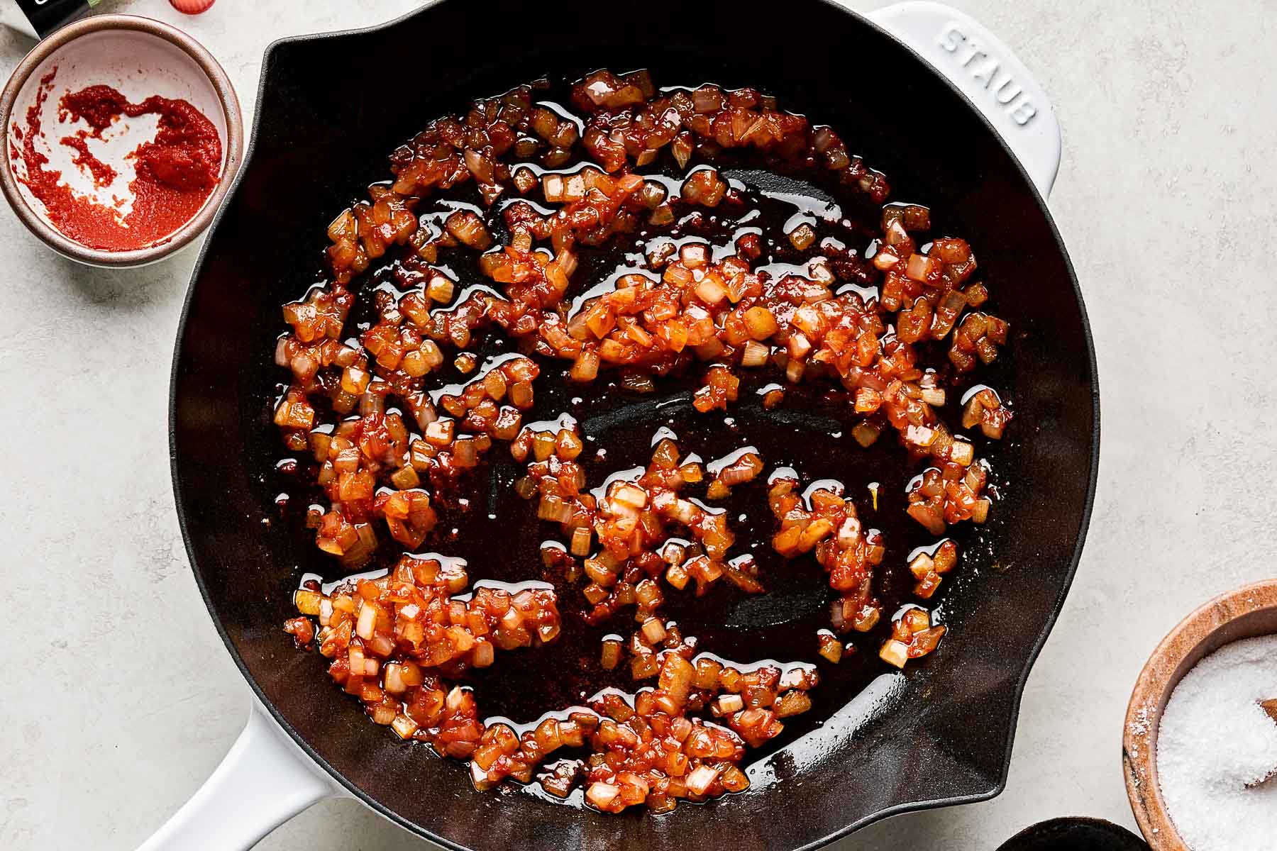An overhead shot of diced onion softened in a cast iron skillet atop a white marbled surface. Small dishes of tomato paste and salt sit alongside the skillet.