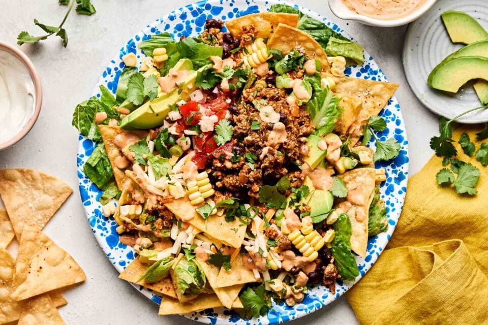 An overhead shot of ground chicken taco salad on a blue and white patterned plate atop a white marbled surface. The plate is surrounded by a yellow cloth napkin and dishes of sour cream, tortilla chips, chipotle ranch dressing and avocado slices.