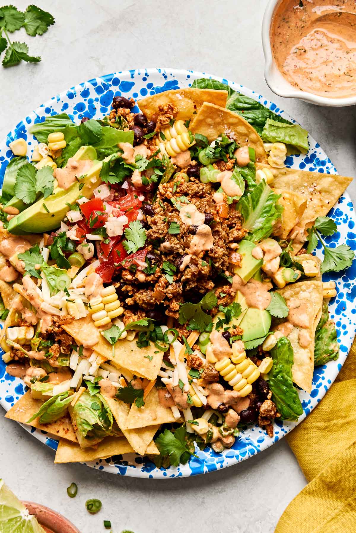 An overhead shot of ground chicken taco salad on a blue and white patterned plate atop a white marbled surface. The plate is surrounded by a yellow cloth napkin and dishes of chipotle ranch dressing and lime wedges.