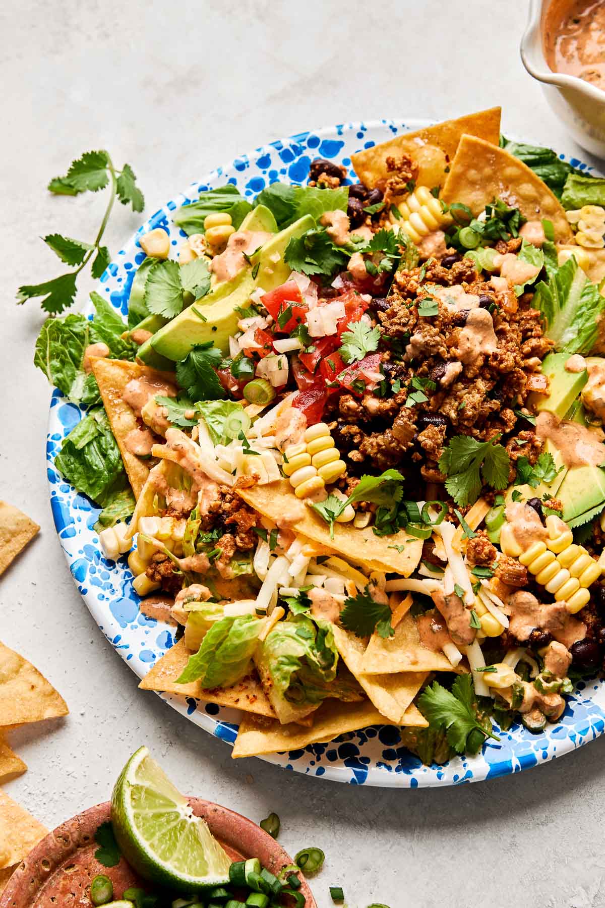 A close-up overhead shot of ground chicken taco salad on a blue and white patterned plate atop a white marbled surface. The plate is surrounded by a bowl of chipotle ranch dressing, tortilla chips and a small dish of lime wedges.