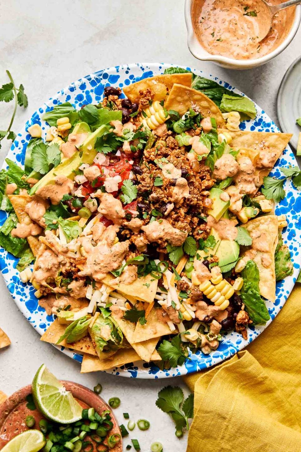 An overhead shot of ground chicken taco salad on a blue and white patterned plate atop a white marbled surface. The plate is surrounded by a yellow cloth napkin and dishes of chipotle ranch dressing and lime wedges.