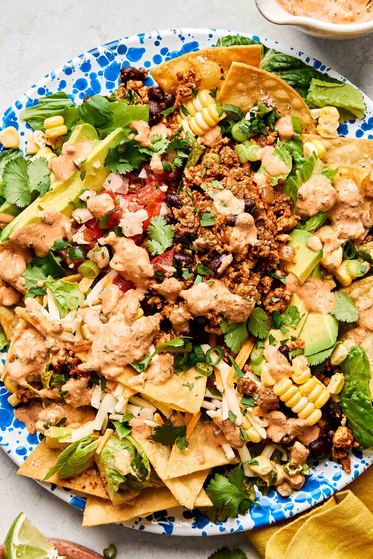 An overhead shot of ground chicken taco salad on a blue and white patterned plate atop a white marbled surface. The plate is surrounded by a yellow cloth napkin and dishes of chipotle ranch dressing and lime wedges.