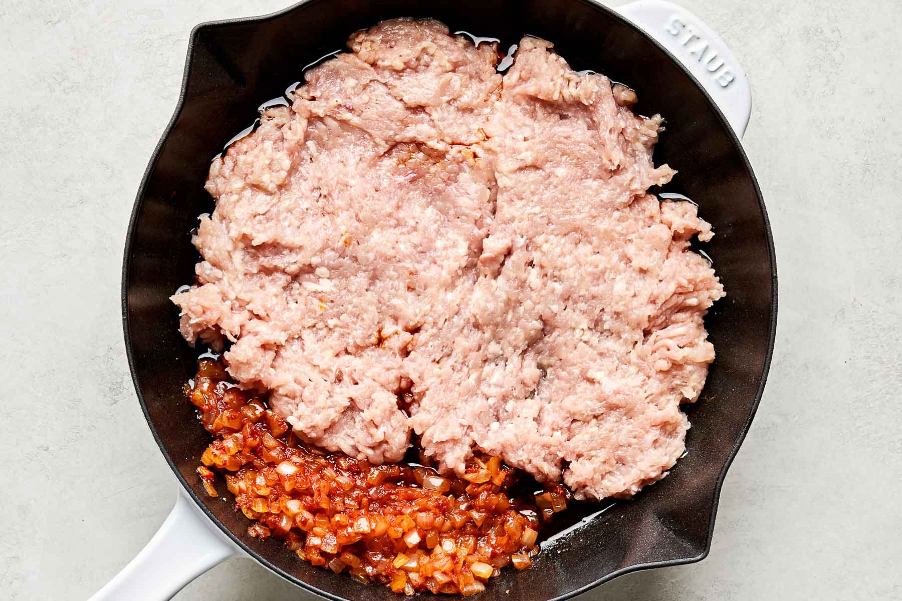 An overhead shot of raw ground chicken and softened onions in a cast iron skillet atop a white marbled surface.