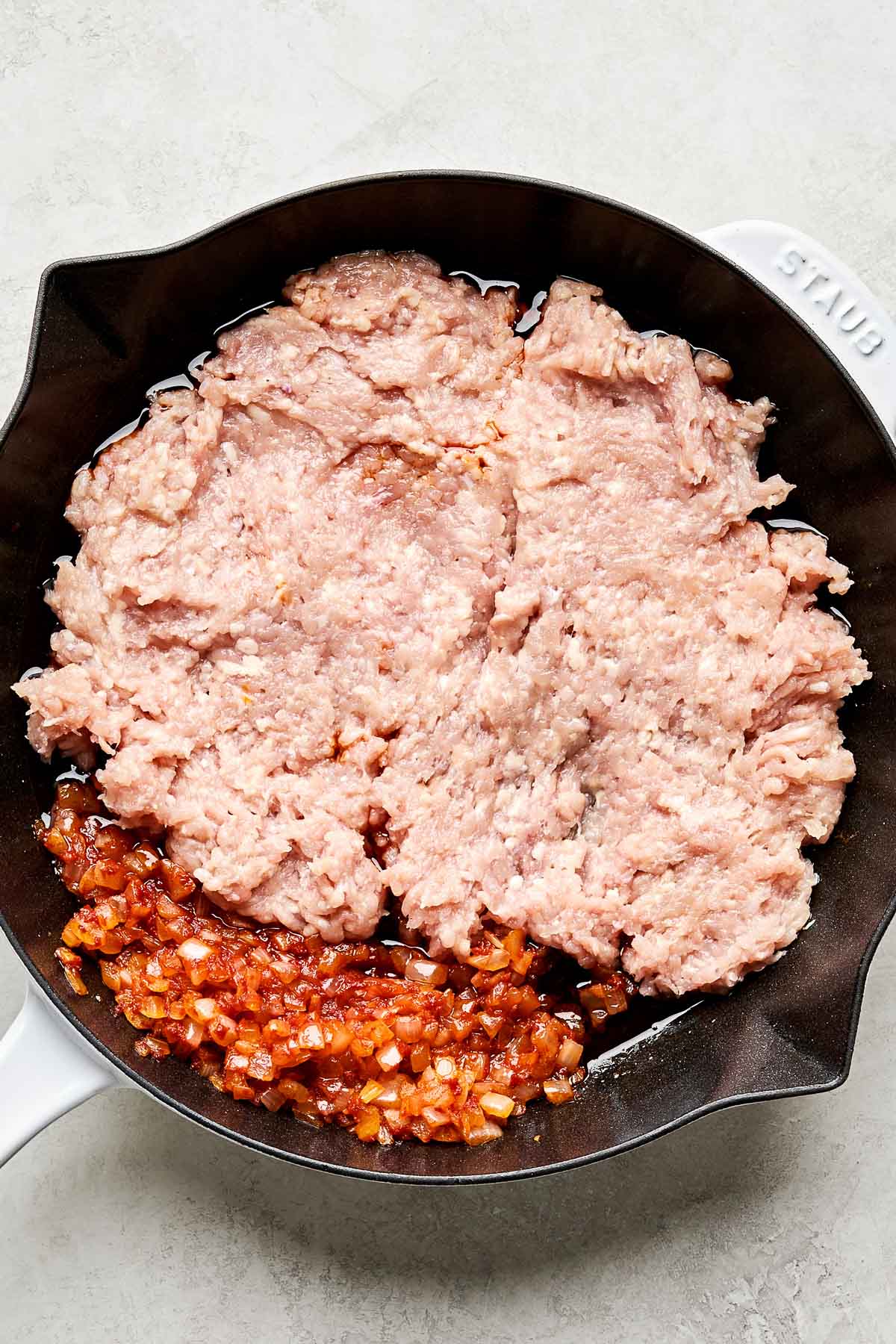 An overhead shot of raw ground chicken and softened onions in a cast iron skillet atop a white marbled surface.