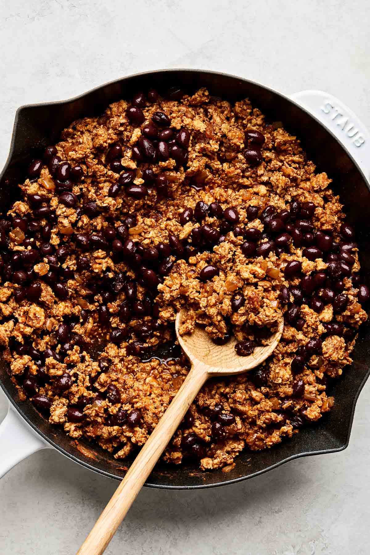 An overhead shot of cooked ground chicken taco meat and black beans with a wooden spoon in a cast iron skillet atop a white marbled surface.
