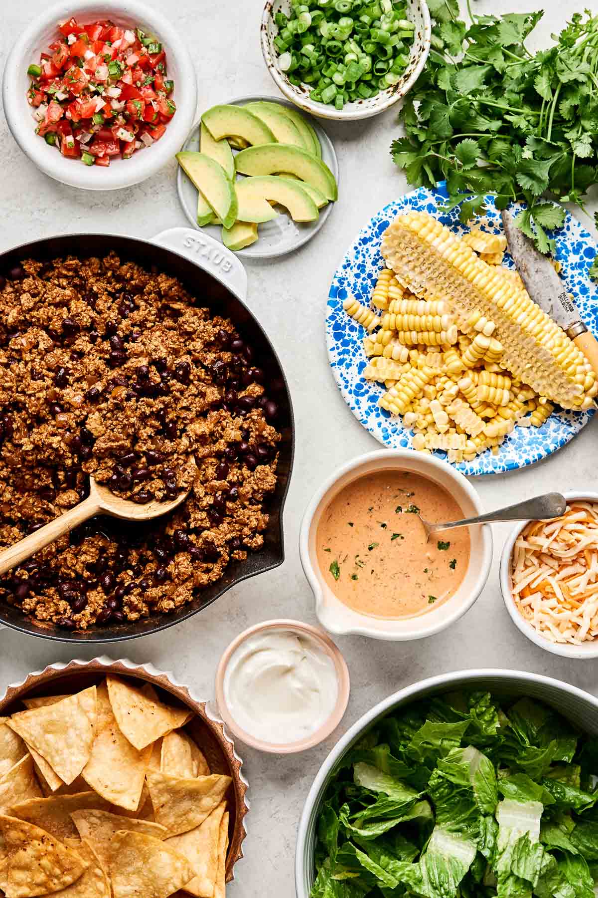 An overhead shot of ingredients displayed on a white marbled surface: a skillet of ground chicken taco meat and dishes of pico de gallo, lettuce, sweet corn, sliced avocado, green onions, shredded cheese, tortilla chips, sour cream, and chipotle ranch.