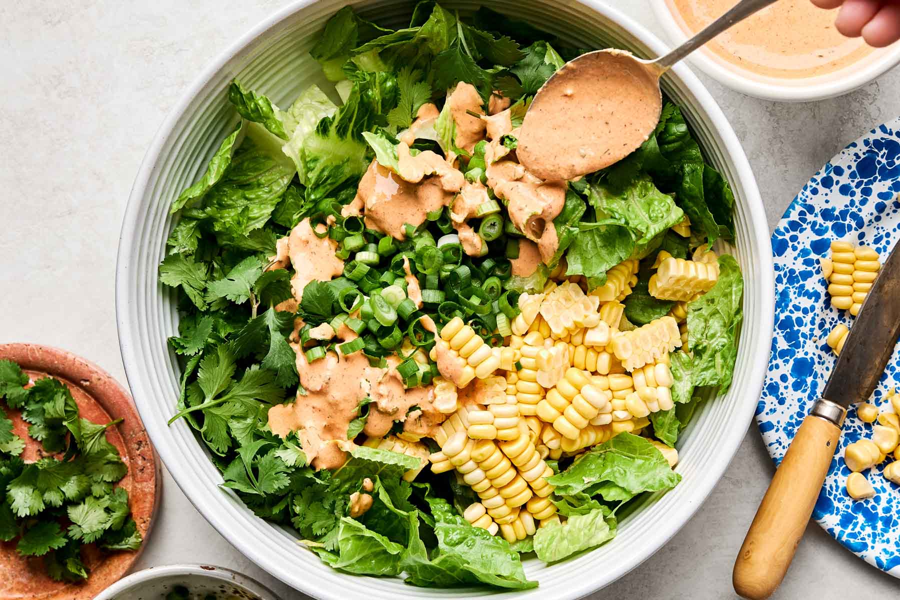 An overhead shot of a spoon drizzling chipotle ranch dressing over a bowl of lettuce, chopped green onions and sweet corn. The bowl sits on a white surface alongside dishes of cilantro, corn, and chipotle ranch.