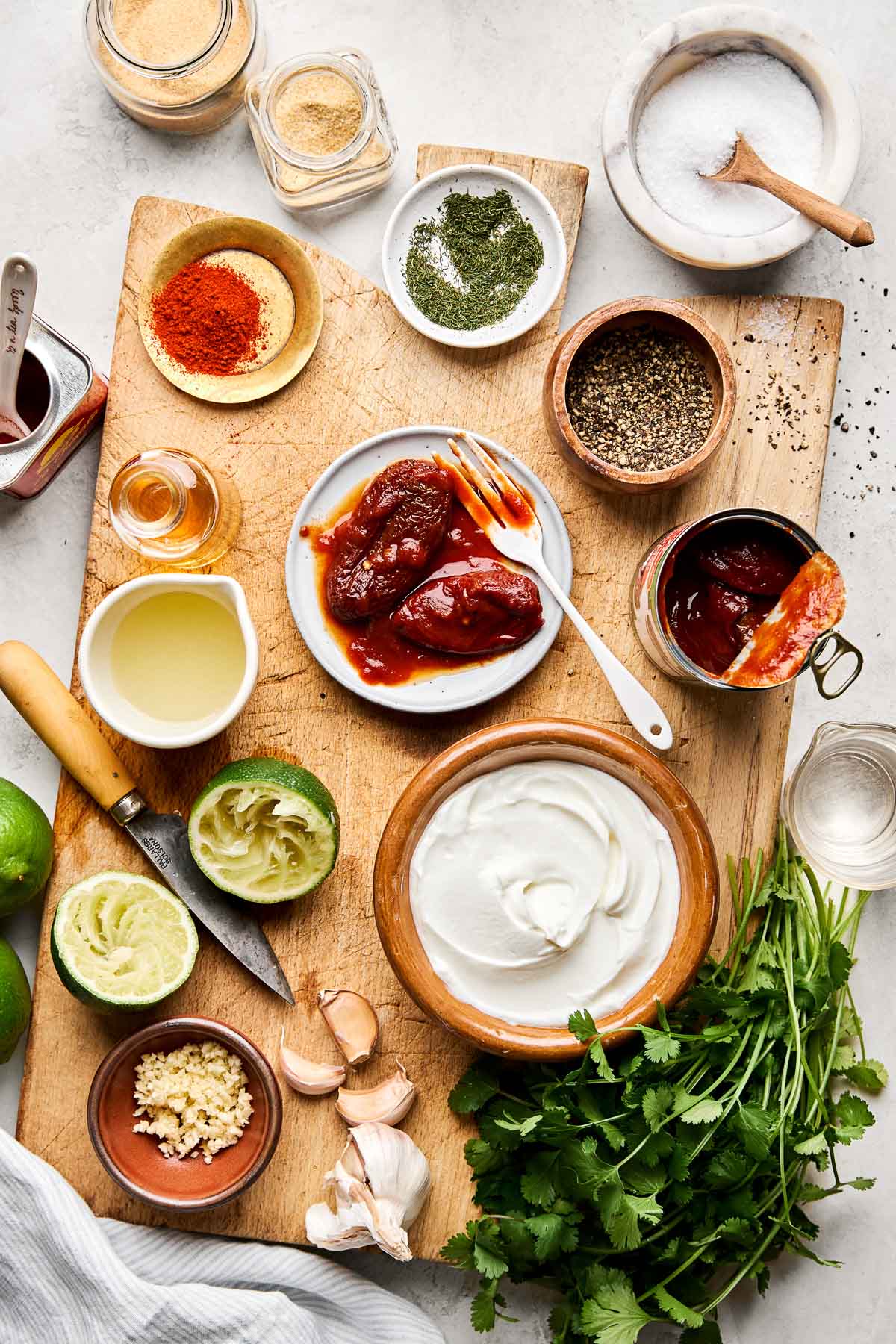 An overhead shot of ingredients displayed in various dishes on a wooden board atop a white marbled surface: Greek yogurt, limes and lime juice, garlic, salt, pepper, chipotle peppers, cilantro, dried dill, garlic powder, and paprika.