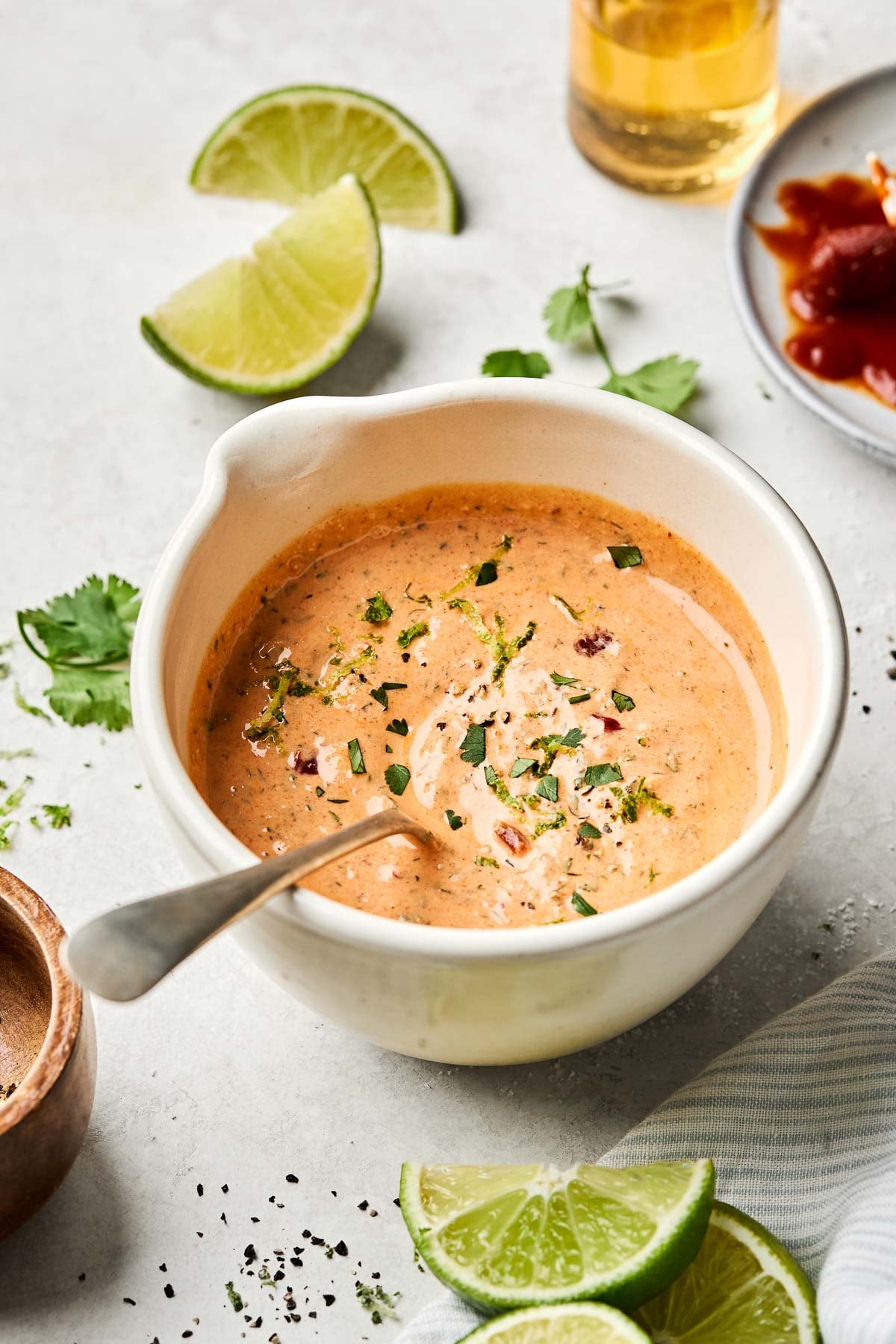 An angled shot of chipotle ranch dressing in a white bowl on a white marbled surface. Lime wedges, cilantro leaves, and dishes of chipotle peppers and ground black pepper surround the bowl.