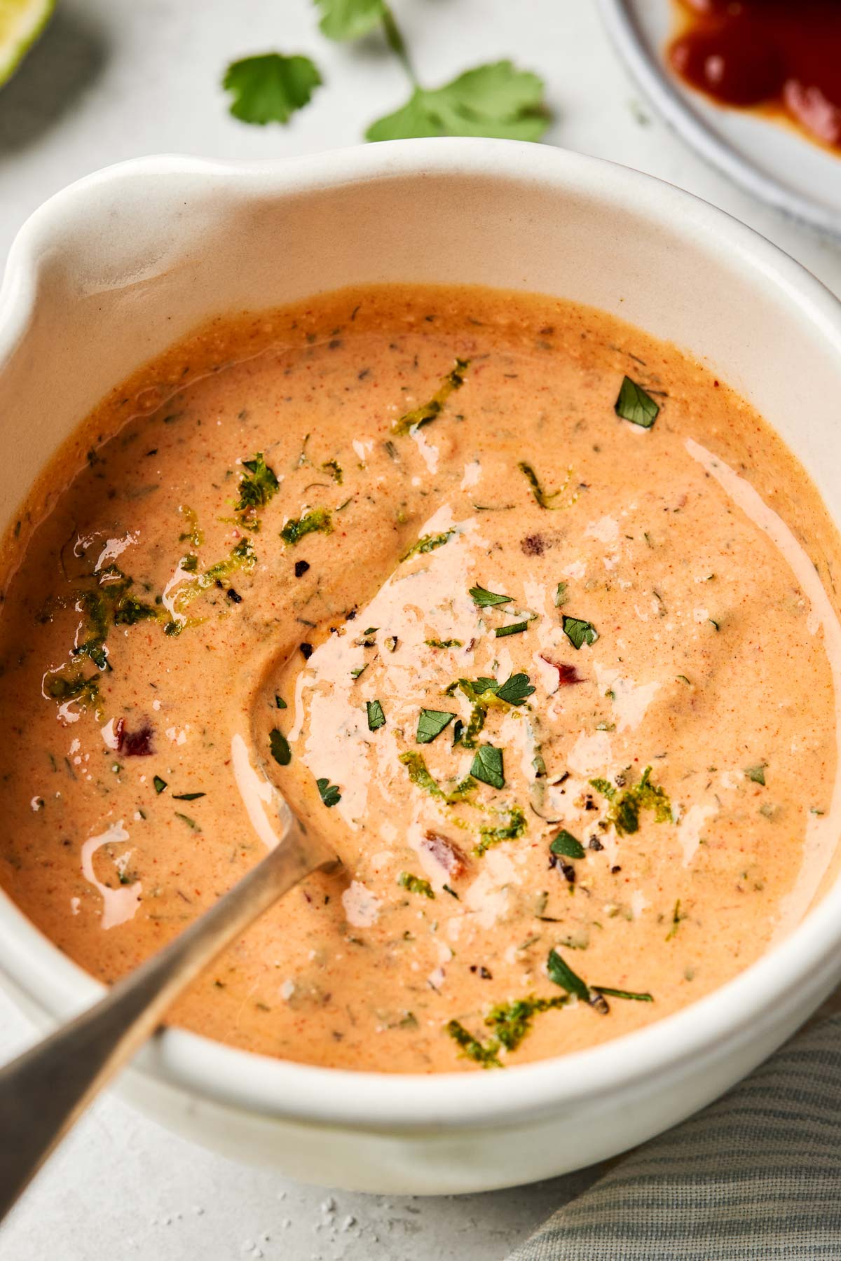 A close-up angled shot of Greek yogurt chipotle ranch in a white bowl atop a white marbled surface. A lime wedge, cilantro leaves and a plate of chipotle peppers can be seen in the background.