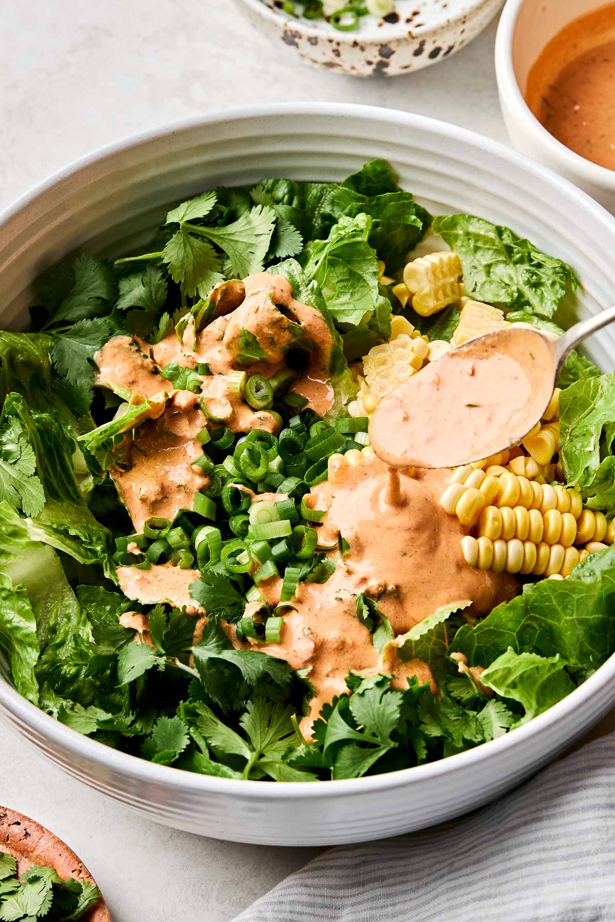 An angled overhead shot of a spoon drizzling chipotle ranch dressing over lettuce, green onions and sweet corn in a large white bowl atop a white marbled surface. Dishes of cilantro, chopped green onions and ranch surround the bowl.