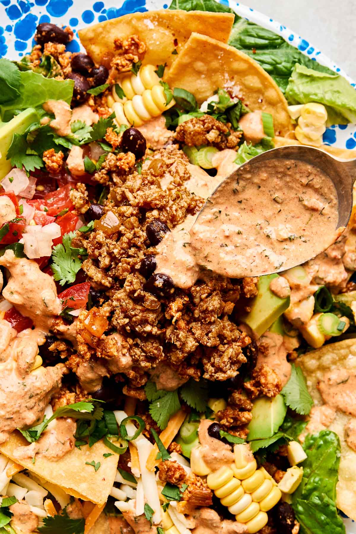 An overhead shot of a spoon drizzling chipotle ranch dressing over a taco salad in a blue and white patterned bowl.