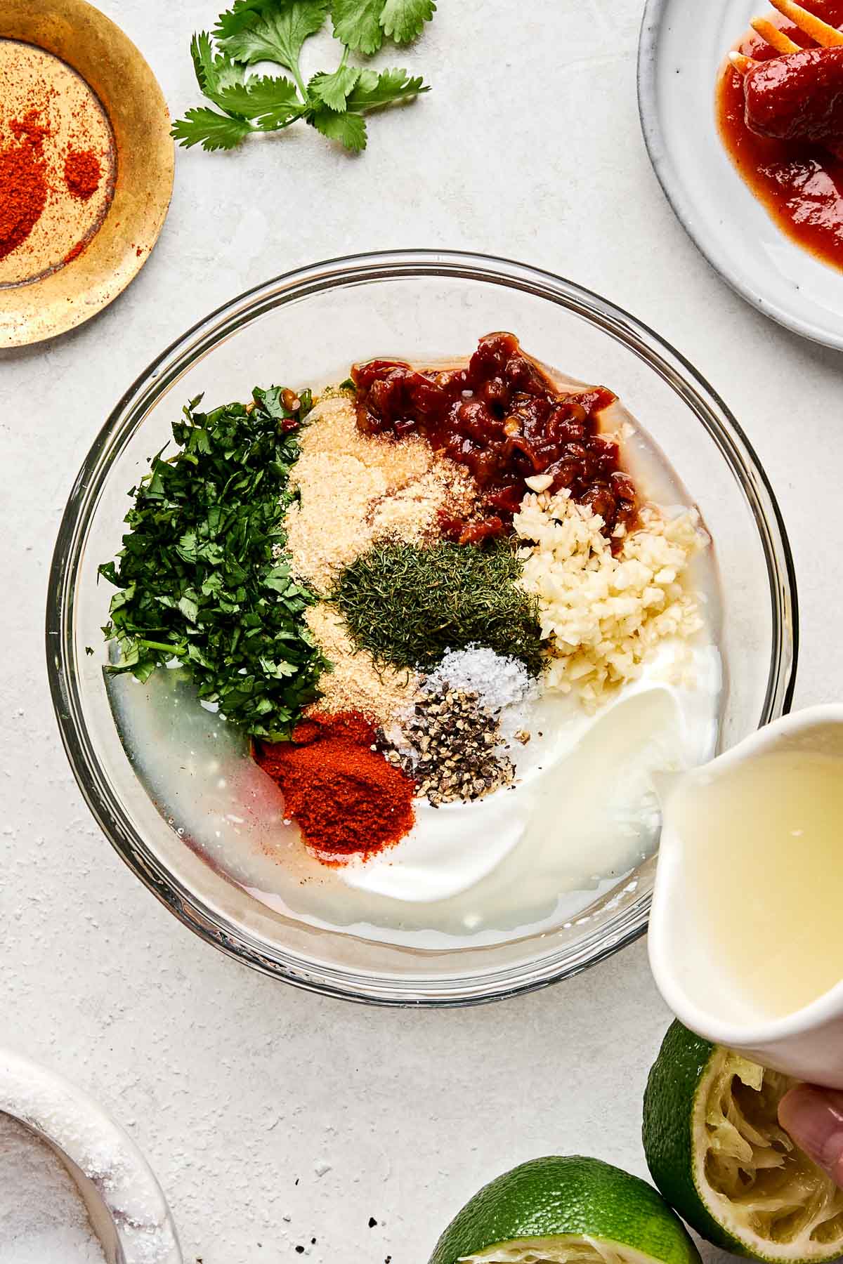 An overhead shot of lime juice being poured into a glass bowl of Greek yogurt, garlic, salt, pepper, chipotle peppers, cilantro, dried dill, garlic powder, and paprika. The bowl sits on a white marbled surface surrounded by dishes of paprika, chipotle peppers, and juiced lime halves.