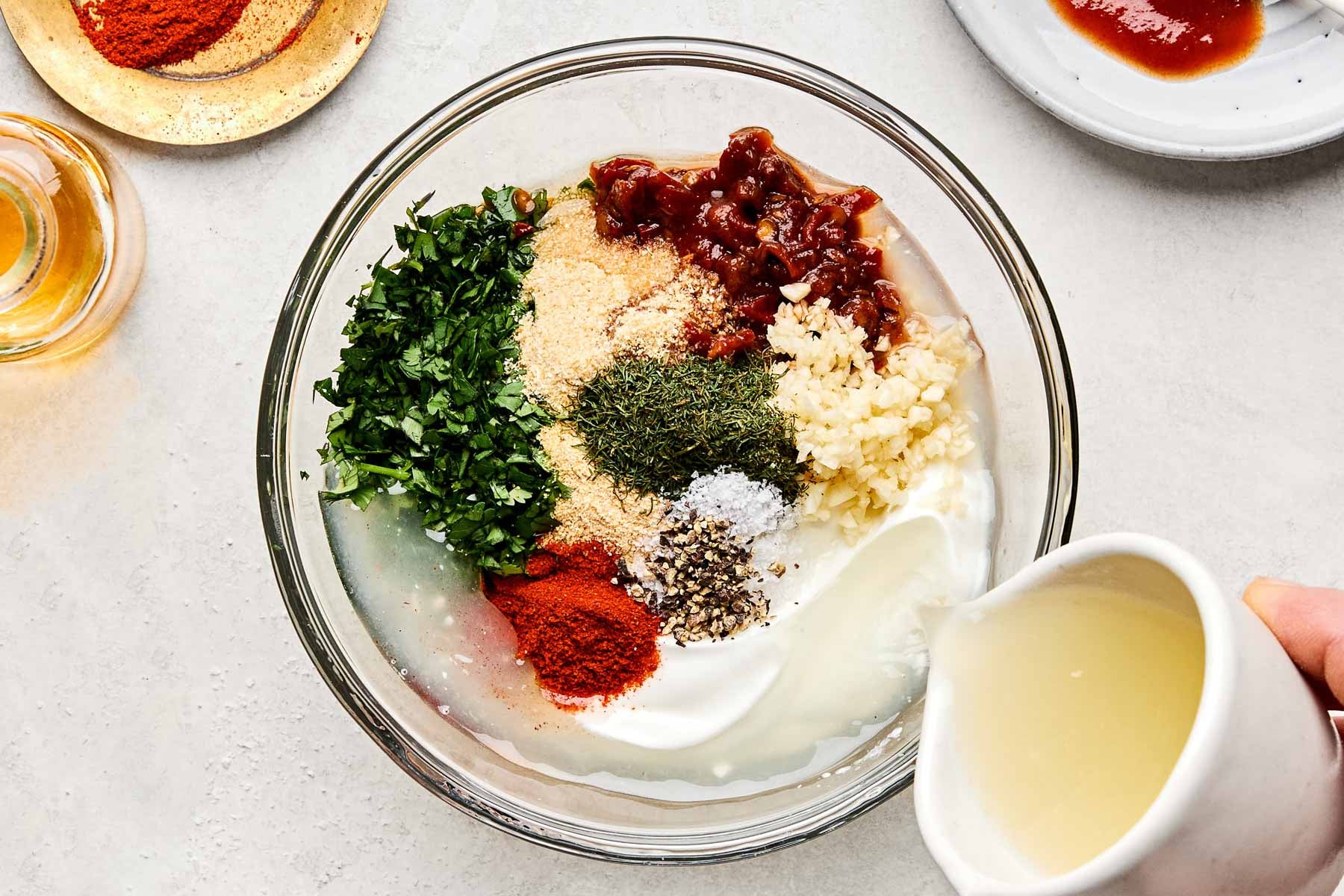 An overhead shot of lime juice being poured into a glass bowl of Greek yogurt, garlic, salt, pepper, chipotle peppers, cilantro, dried dill, garlic powder, and paprika. The bowl sits on a white marbled surface surrounded by dishes of paprika, chipotle peppers, and juiced lime halves.
