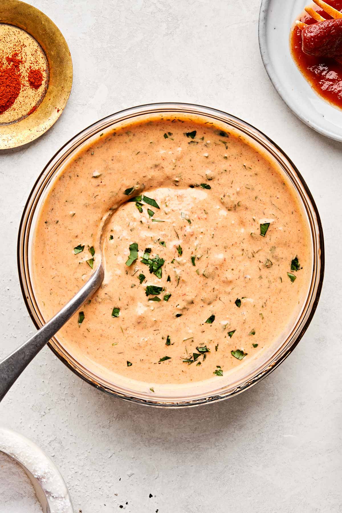 An overhead shot of chipotle ranch dressing with a spoon in a glass bowl atop a white marbled surface. Dishes of salt, paprika and chipotle peppers sit alongside the bowl.
