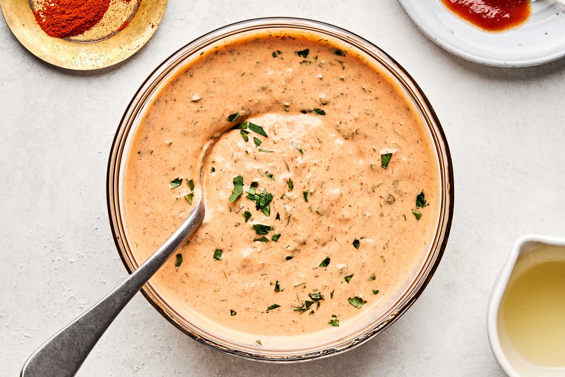 An overhead shot of chipotle ranch dressing with a spoon in a glass bowl atop a white marbled surface. Dishes of salt, paprika and chipotle peppers sit alongside the bowl.
