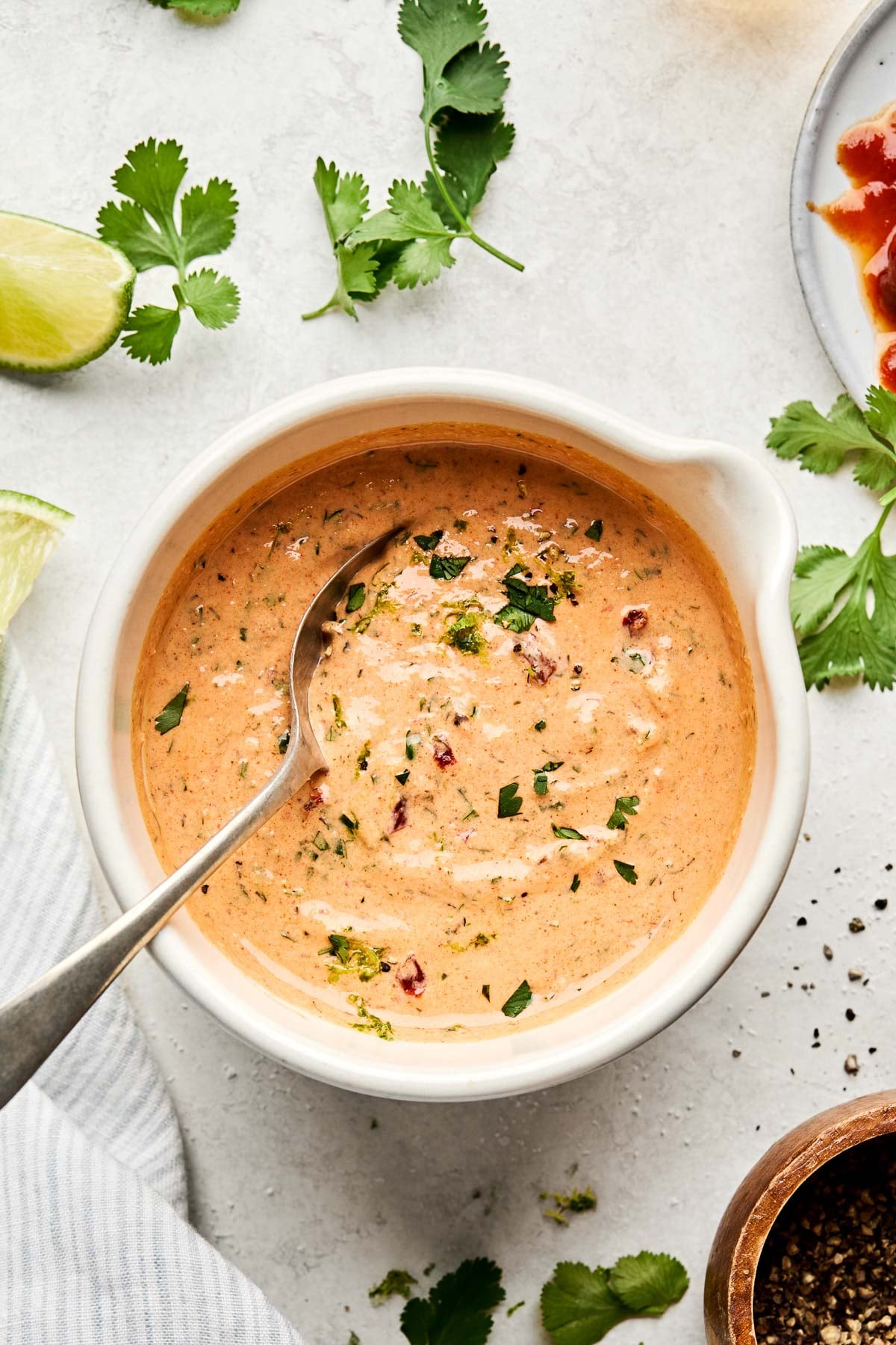 An overhead shot of Greek yogurt chipotle ranch dressing in a white dish on a white marbled surface. The dish is surrounded by cilantro leaves, lime wedges, and dishes of chipotle peppers and ground black pepper.