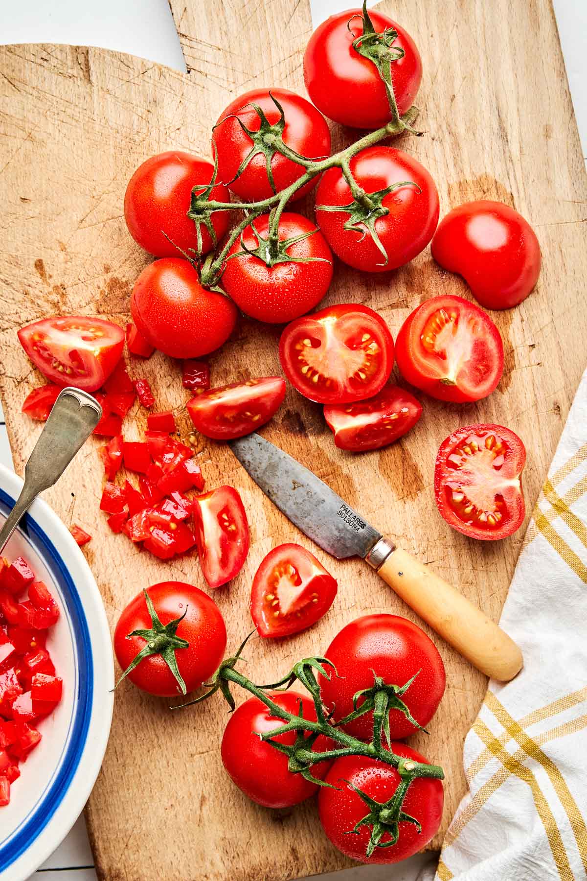 An overhead shot of whole tomatoes on the vine and sliced tomatoes on a wooden cutting board atop a white surface.