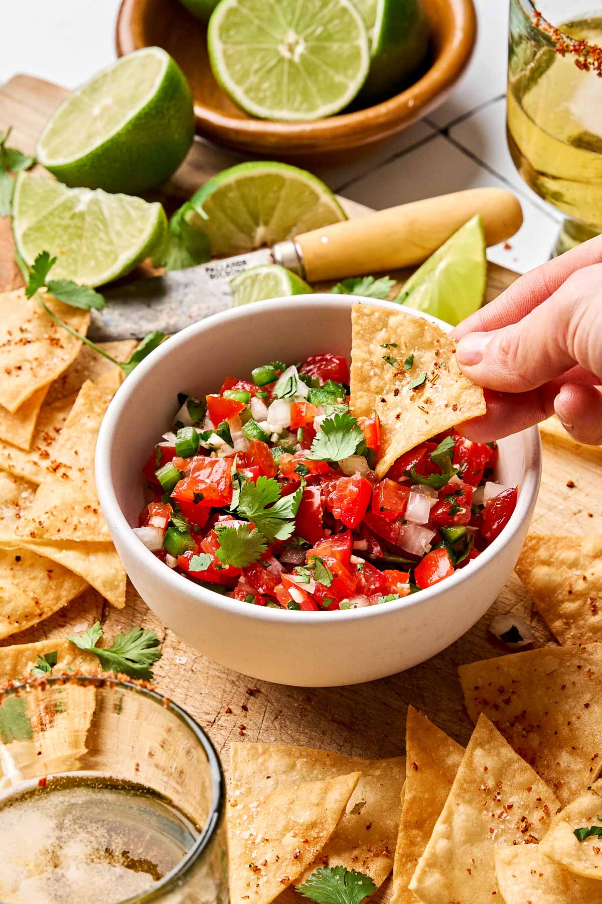 An angled shot of a woman's hand dipping a tortilla chip into a small white bowl of tomato salsa. The bowl sits on a wooden cutting board atop a white tiled surface, surrounded by tortilla chips, margaritas, and lime halves and wedges.