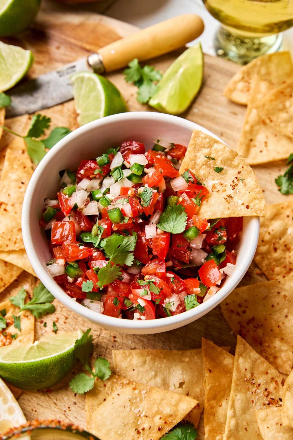 An angled shot of a tortilla chip in a small white bowl of tomato salsa. The bowl sits on a wooden cutting board atop a white tiled surface, surrounded by tortilla chips, margaritas, and lime halves and wedges.