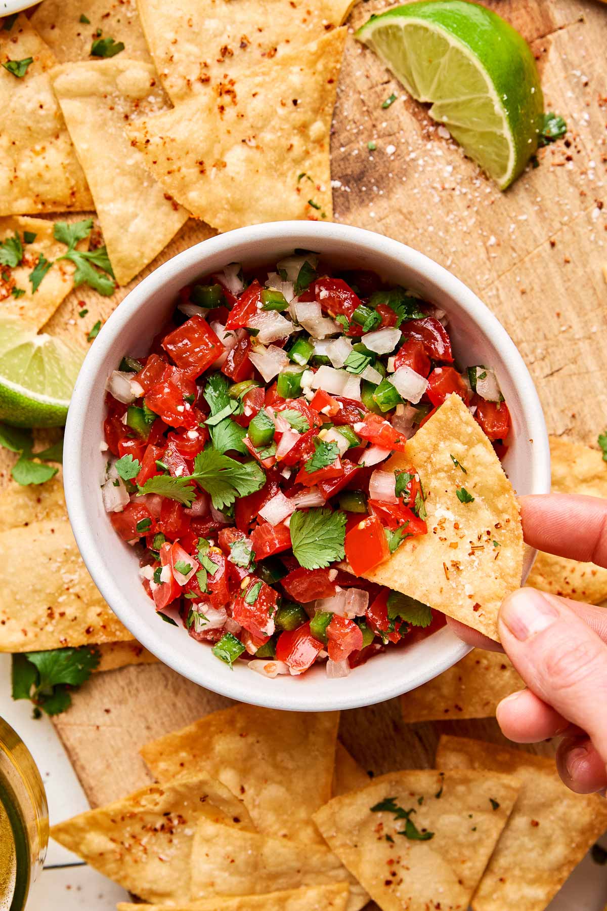 An overhead shot of a woman's hand dipping a tortilla chip into a small white bowl of tomato salsa. The bowl sits on a wooden cutting board, surrounded by tortilla chips and lime wedges.