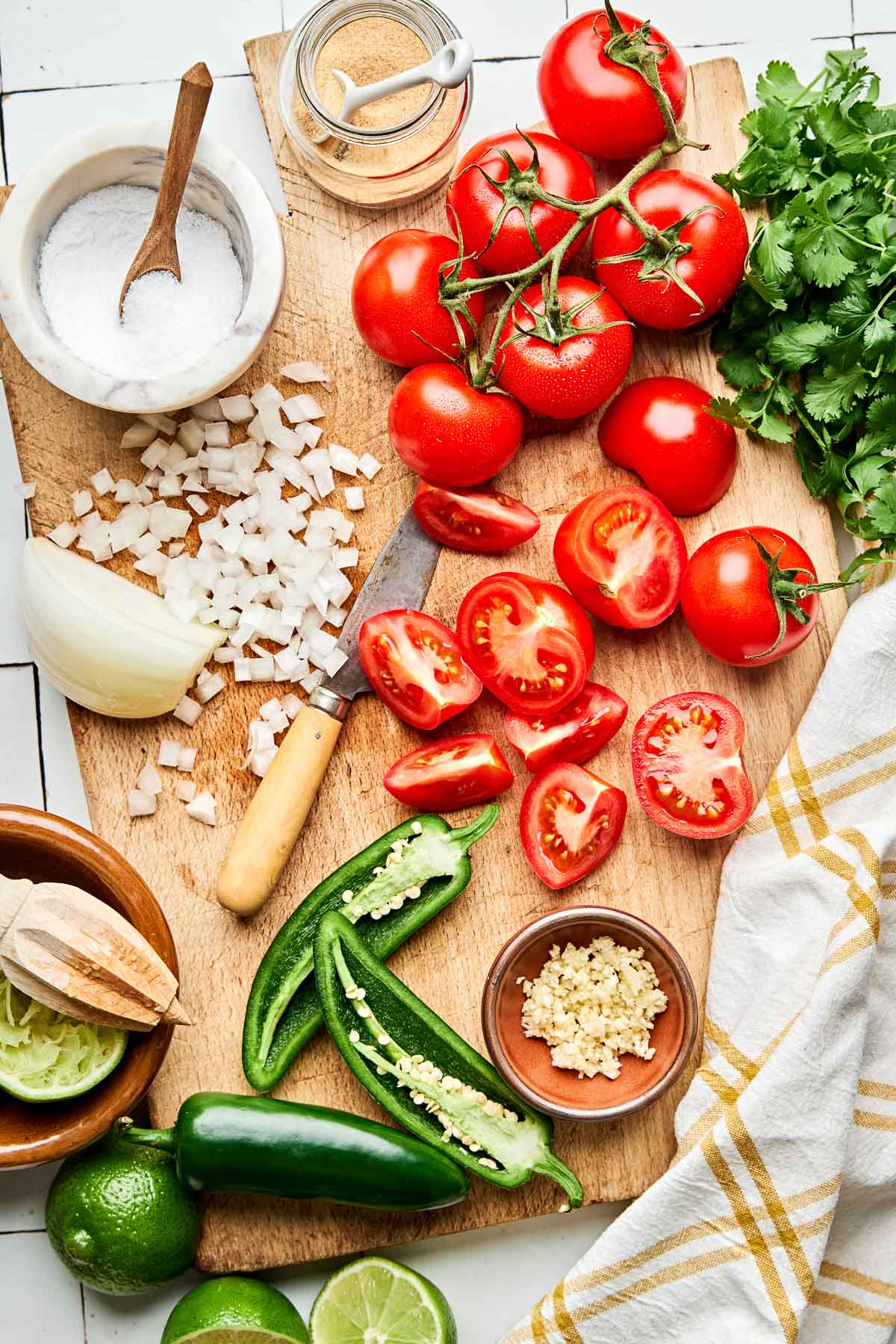 An overhead shot of ingredients displayed on a wooden cutting board atop a white tiled surface: whole and sliced tomatoes, whole and diced onion, cilantro, whole and halved jalapenos, limes, salt, and garlic powder.