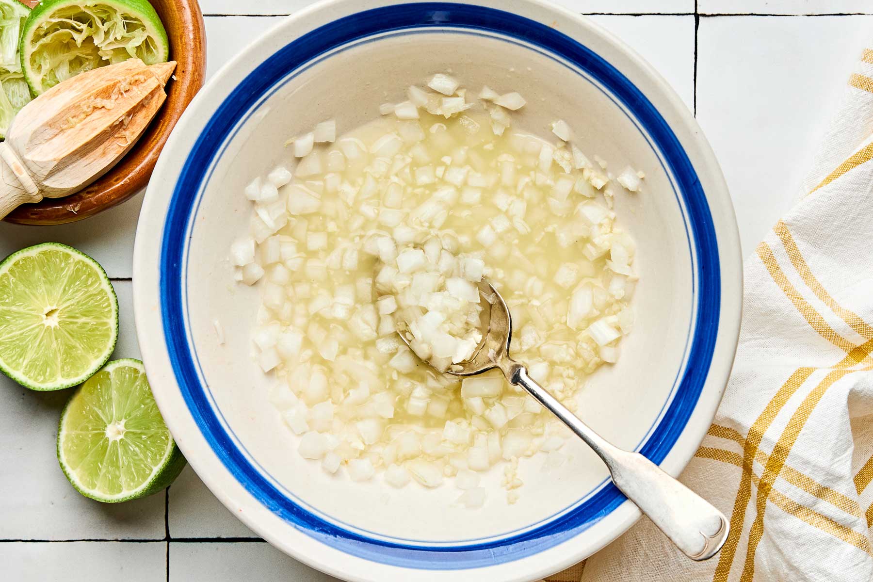 An overhead shot of chopped onions soaking in lime juice in a white bowl with a blue rim. The bowl sits on a white tiled surface alongside halved and juiced limes and a yellow and white cloth napkin.