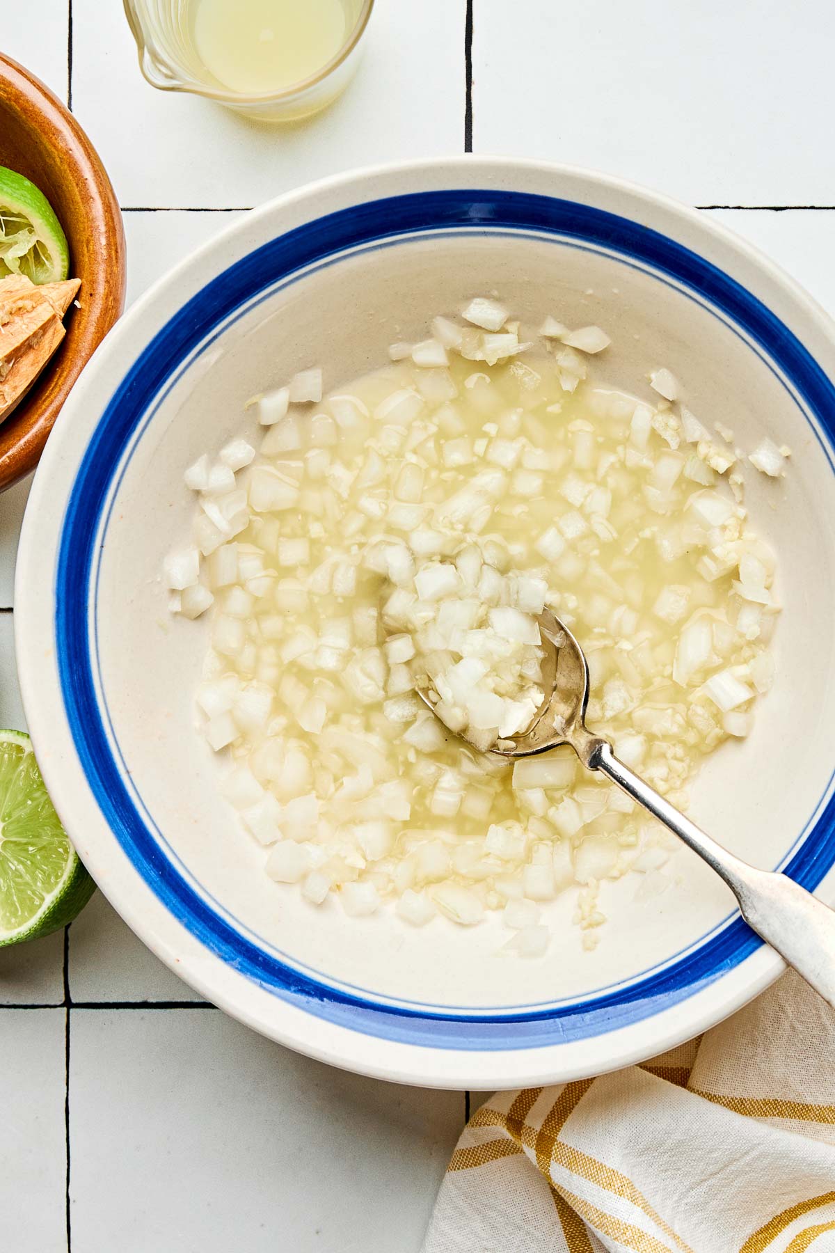 An overhead shot of chopped onions soaking in lime juice in a white bowl with a blue rim. The bowl sits on a white tiled surface alongside halved and juiced limes and a yellow and white cloth napkin.
