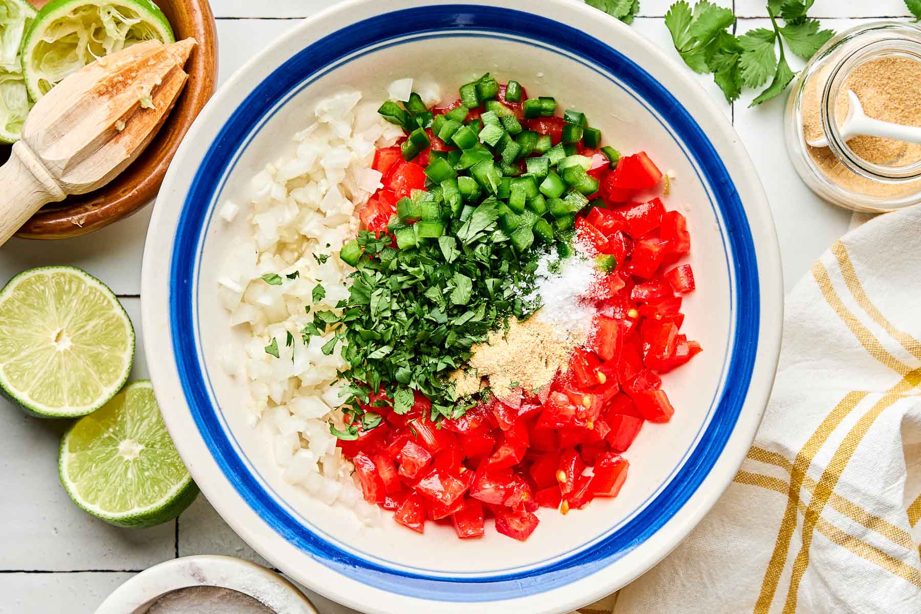 An overhead shot of diced tomatoes, onions, jalapeno, cilantro, garlic powder and salt in a white bowl with a blue rim. The bowl sits on a white tiled surface alongside a white and yellow plaid cloth napkin, halved and juiced limes, cilantro, and a jar of garlic powder.